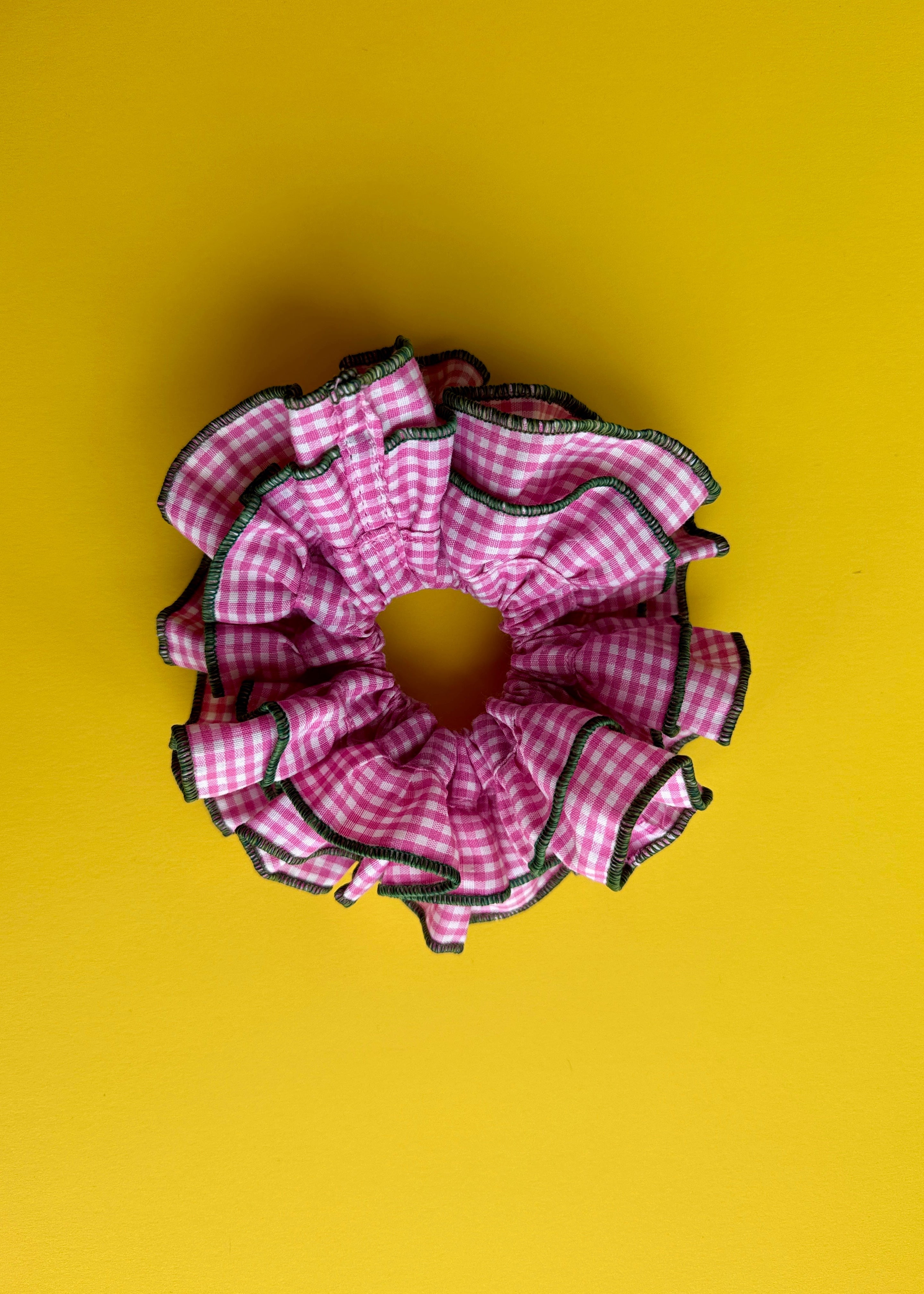 A pink and white gingham ruffled hair scrunchie with black trim, photographed from directly above on a solid yellow background.