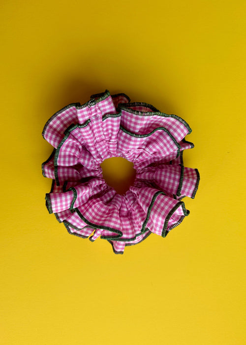 A pink and white gingham ruffled hair scrunchie with black trim, photographed from directly above on a solid yellow background.