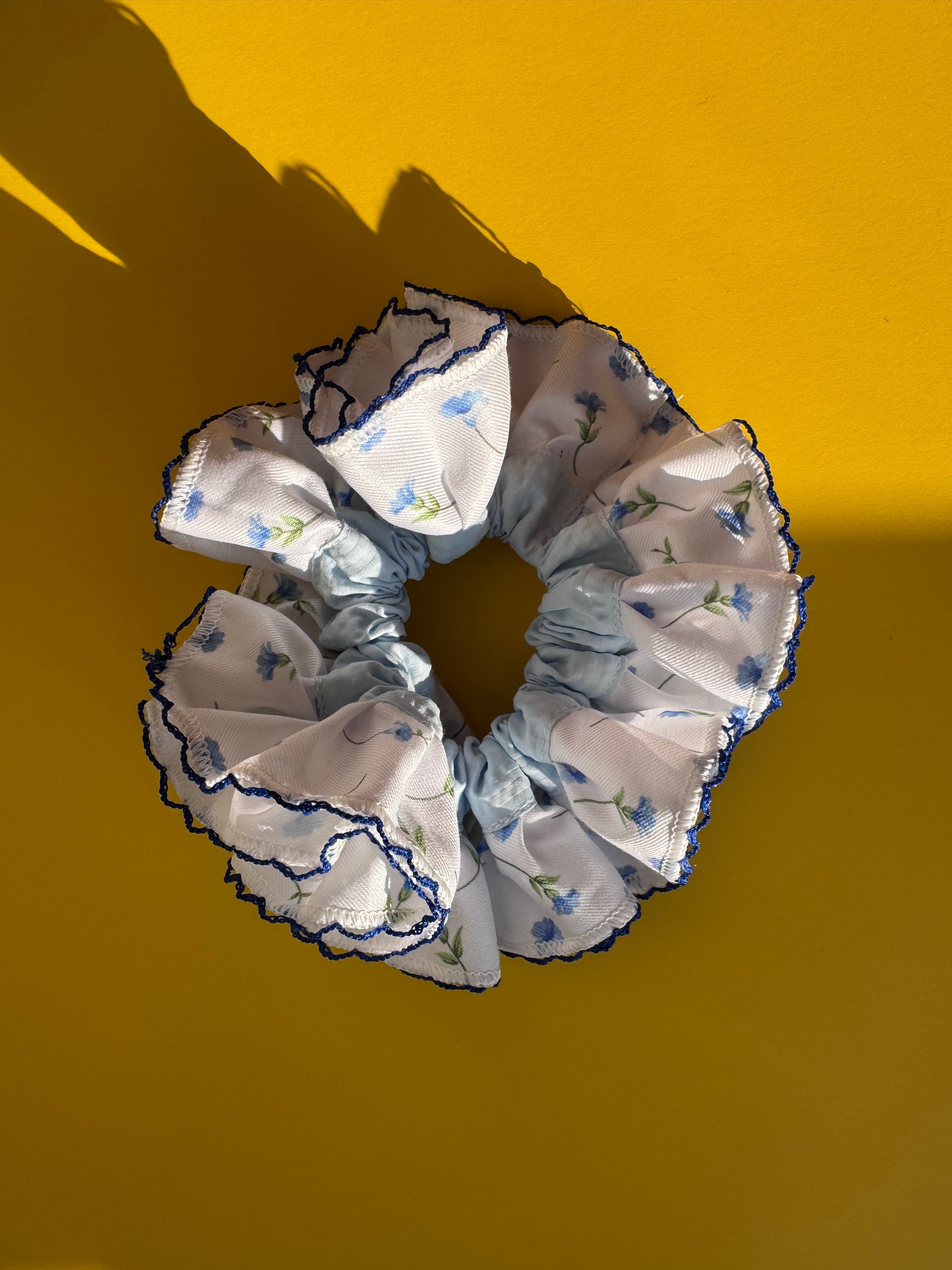 A fabric scrunchie with a white background, light blue floral print, and dark blue ruffled trim, photographed on a solid yellow background with a strong shadow.