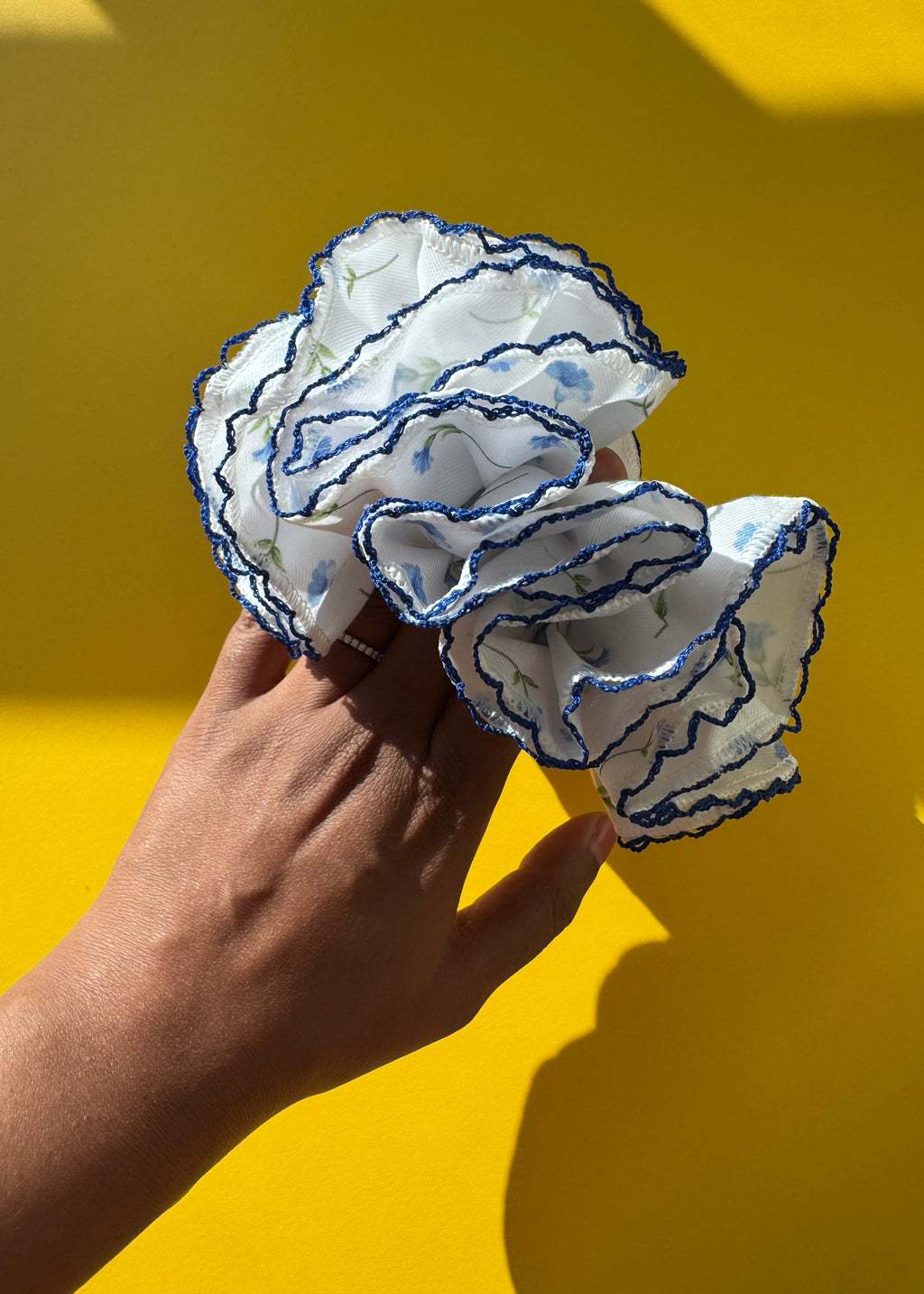A hand holding a white ruffled fabric hair scrunchie with blue embroidered trim and a subtle blue floral pattern, set against a bright yellow background.