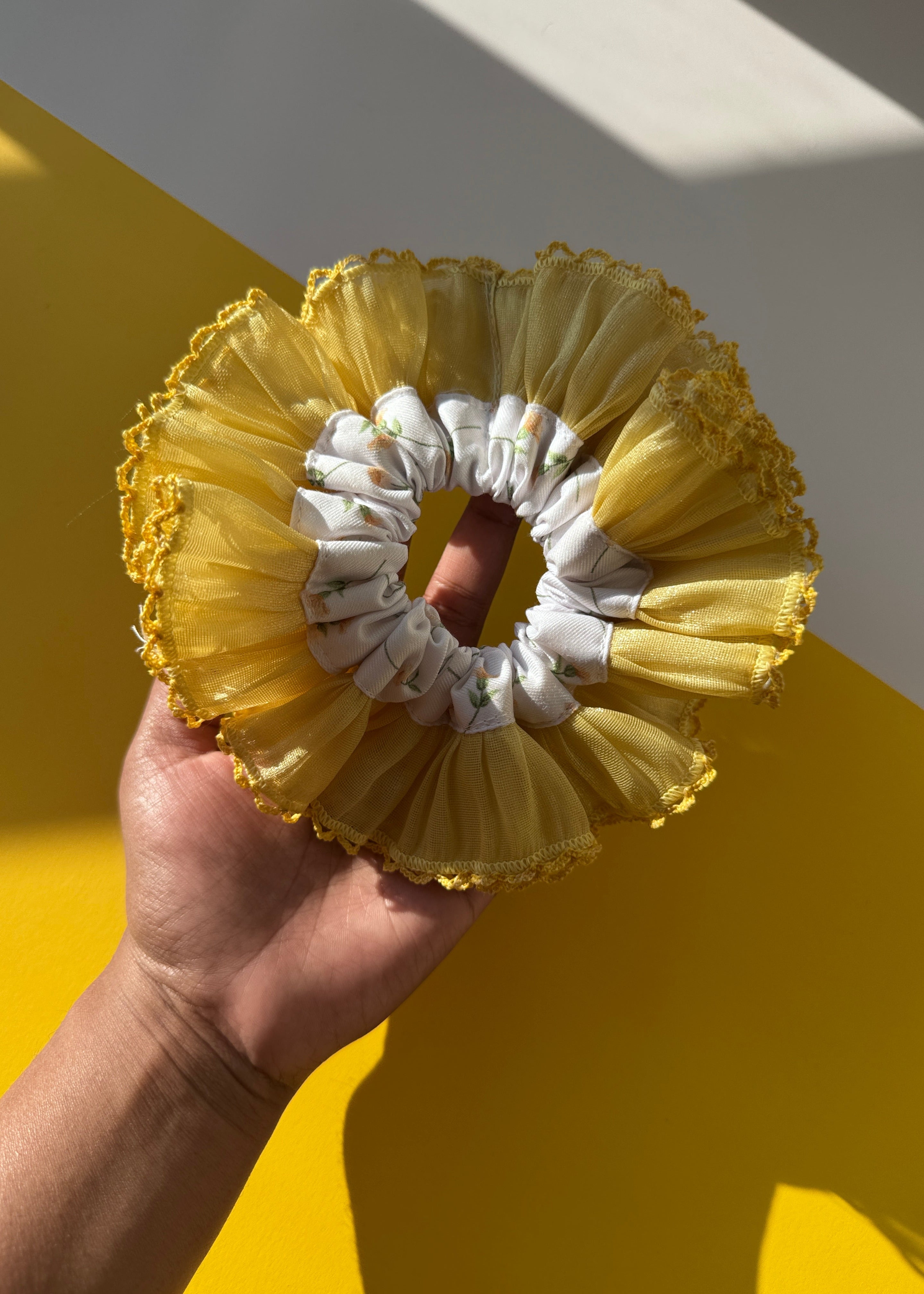 A hand holding a yellow and white ruffled hair scrunchie against a yellow and white background.