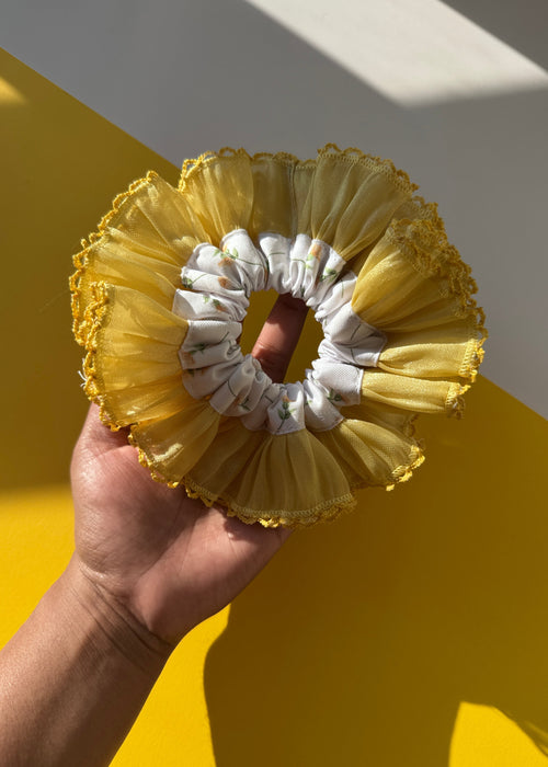 A hand holding a yellow and white ruffled hair scrunchie against a yellow and white background.