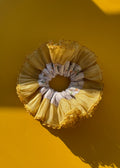 A ruffled fabric hair scrunchie in a pale yellow and white patterned design, displayed against a solid yellow background with a strong shadow.