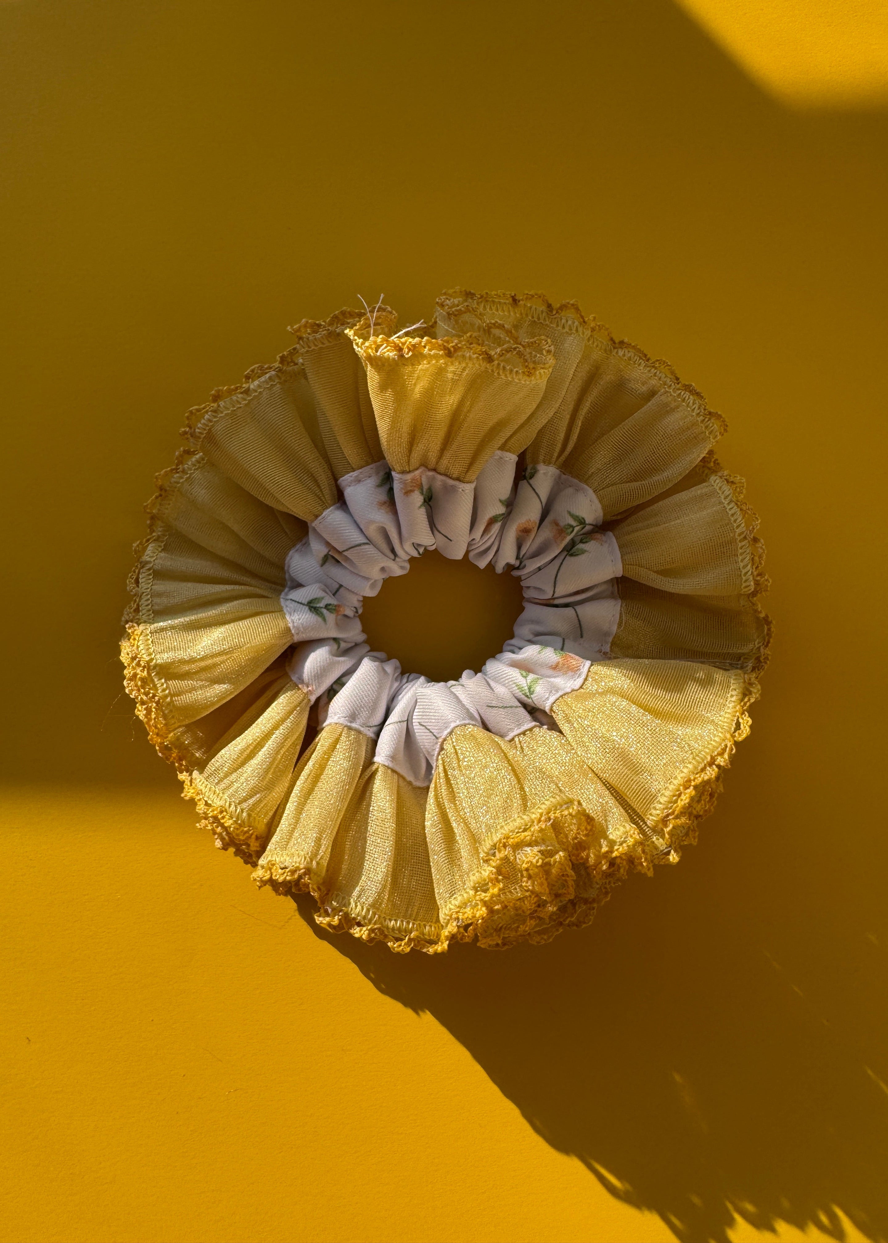A ruffled fabric hair scrunchie in a pale yellow and white patterned design, displayed against a solid yellow background with a strong shadow.