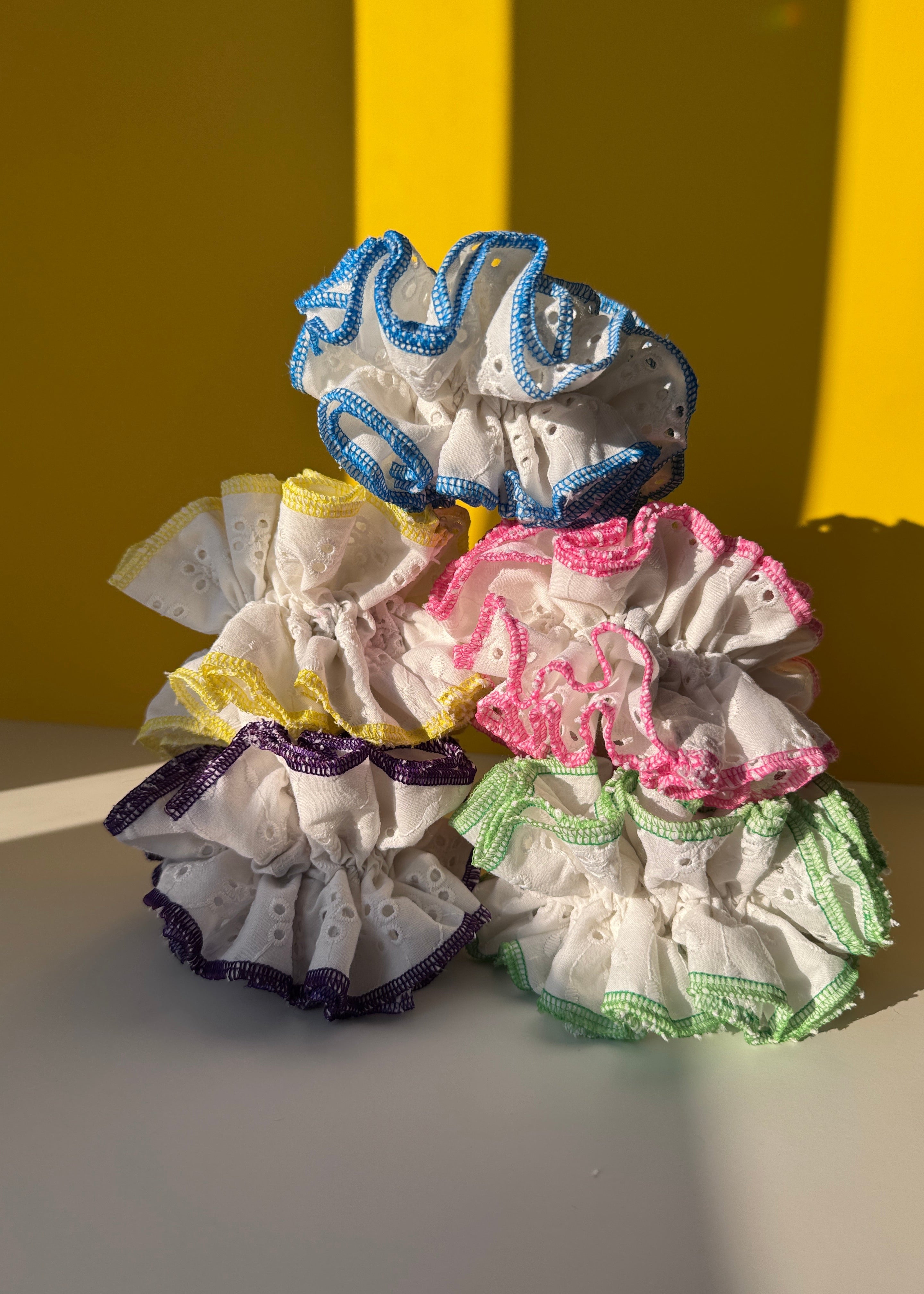 Five eyelet fabric hair scrunchies in assorted colors (blue, pink, green, yellow, and purple) arranged on a split yellow and white background.