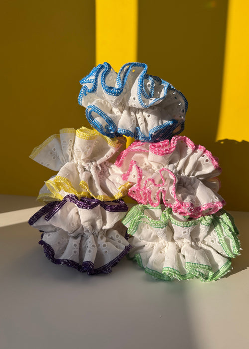 Five eyelet fabric hair scrunchies in assorted colors (blue, pink, green, yellow, and purple) arranged on a split yellow and white background.