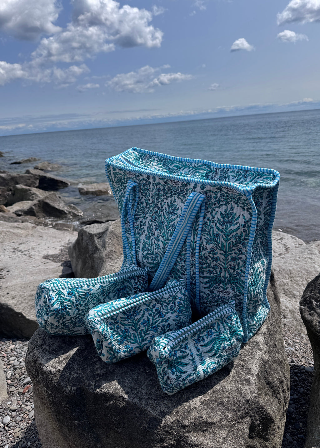 A blue and white patterned knit beach bag and matching pouches are draped over large rocks by the ocean, with water, cloudy sky, and shoreline visible in the background.