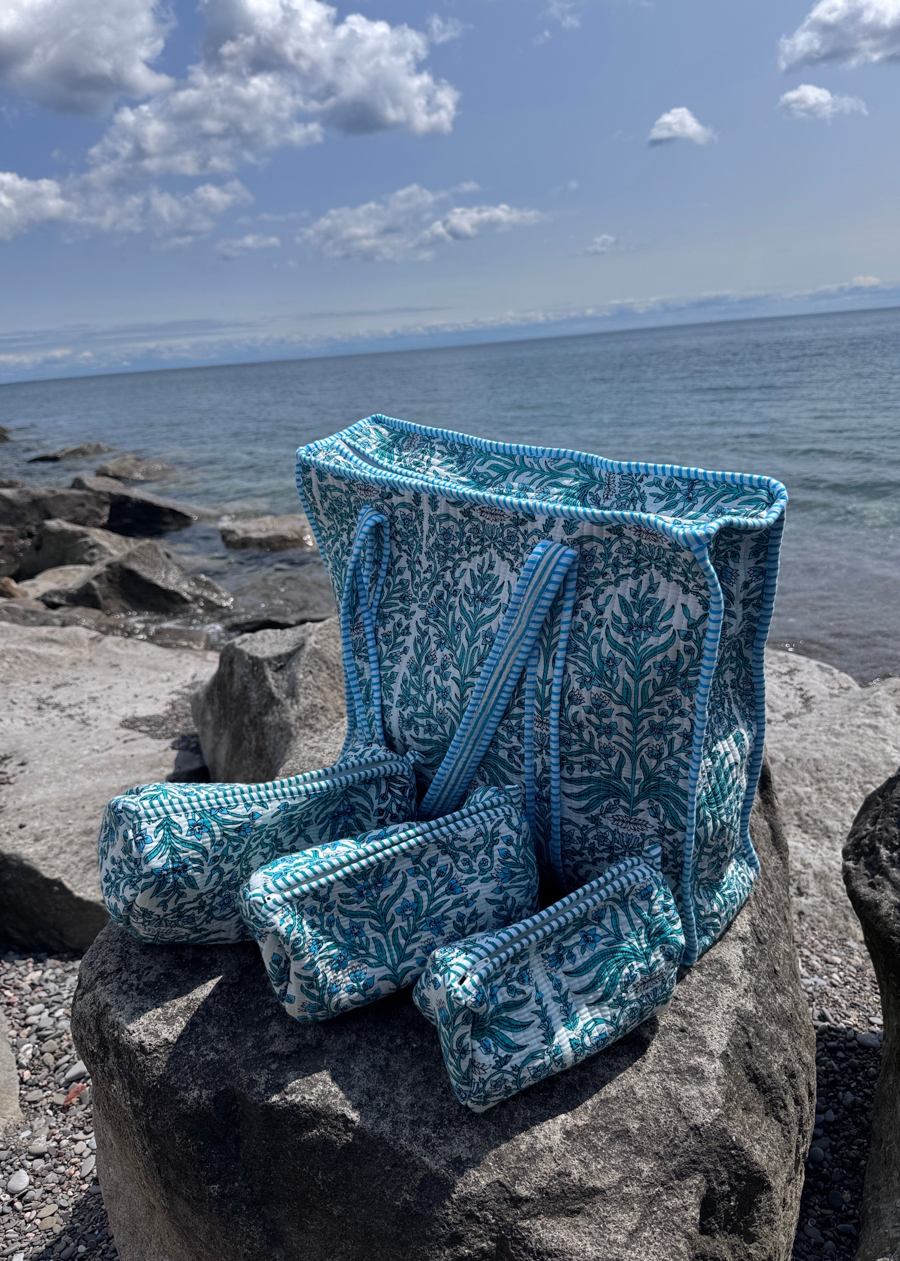 A blue and white patterned knit beach bag and matching pouches are draped over large rocks by the ocean, with water, cloudy sky, and shoreline visible in the background.