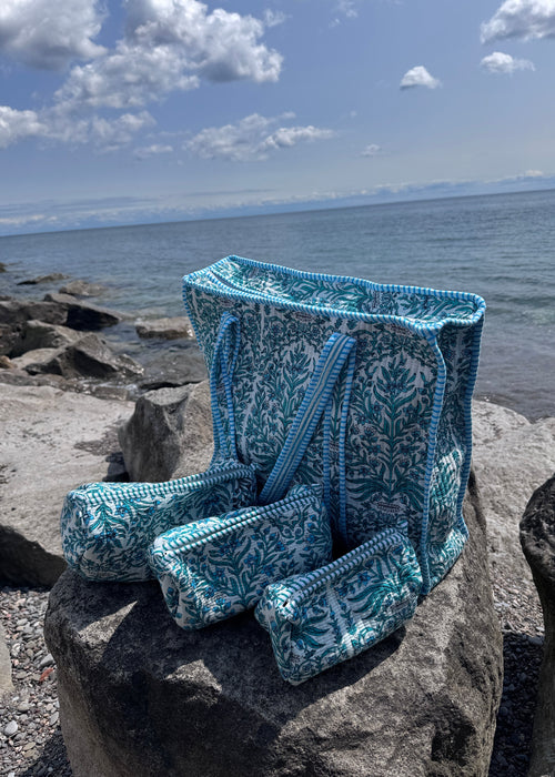 A blue and white patterned knit beach bag and matching pouches are draped over large rocks by the ocean, with water, cloudy sky, and shoreline visible in the background.