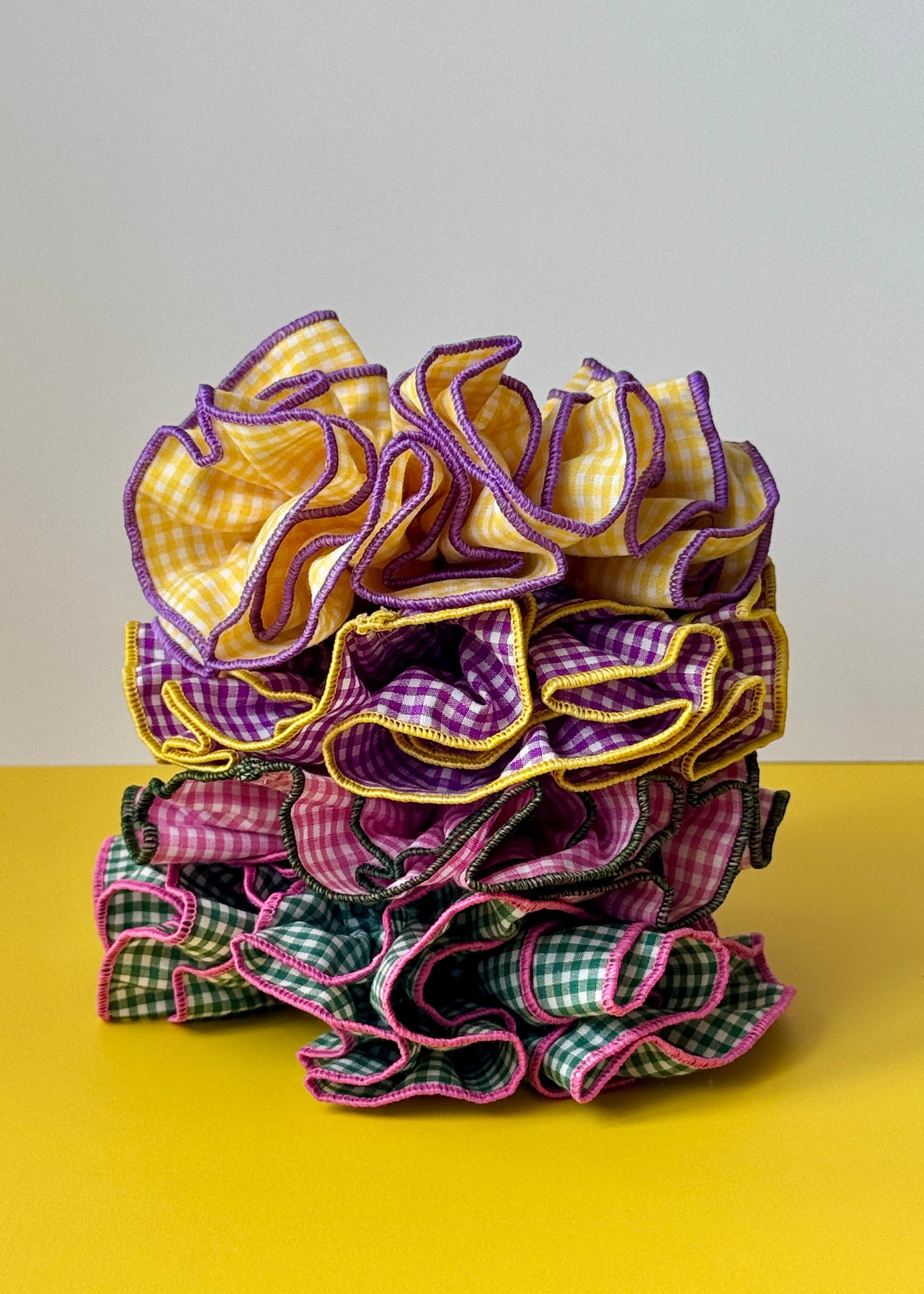 A stack of ruffled fabric hair ties or scrunchies in gingham patterns, featuring purple, yellow, pink, and green colors with contrasting stitched edges, set against a split yellow and gray background.
