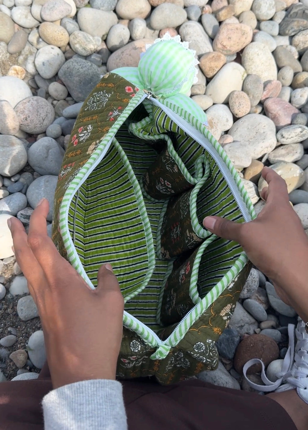 A green and white striped fabric organizer pouch with a floral patterned exterior, shown open with hands holding it on a bed of smooth river stones.