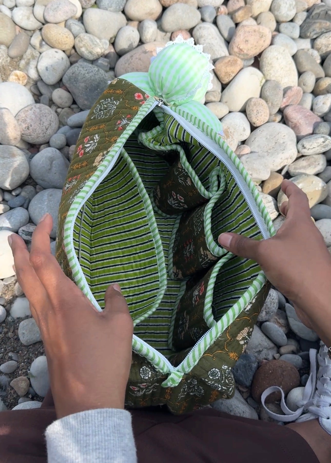 A green and white striped fabric organizer pouch with a floral patterned exterior, shown open with hands holding it on a bed of smooth river stones.