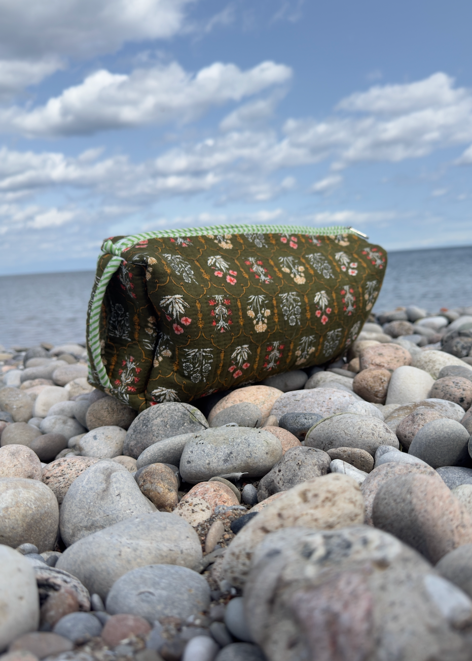A small zippered pouch with a green floral pattern and light green trim sits on a rocky beach with the ocean and cloudy sky in the background.