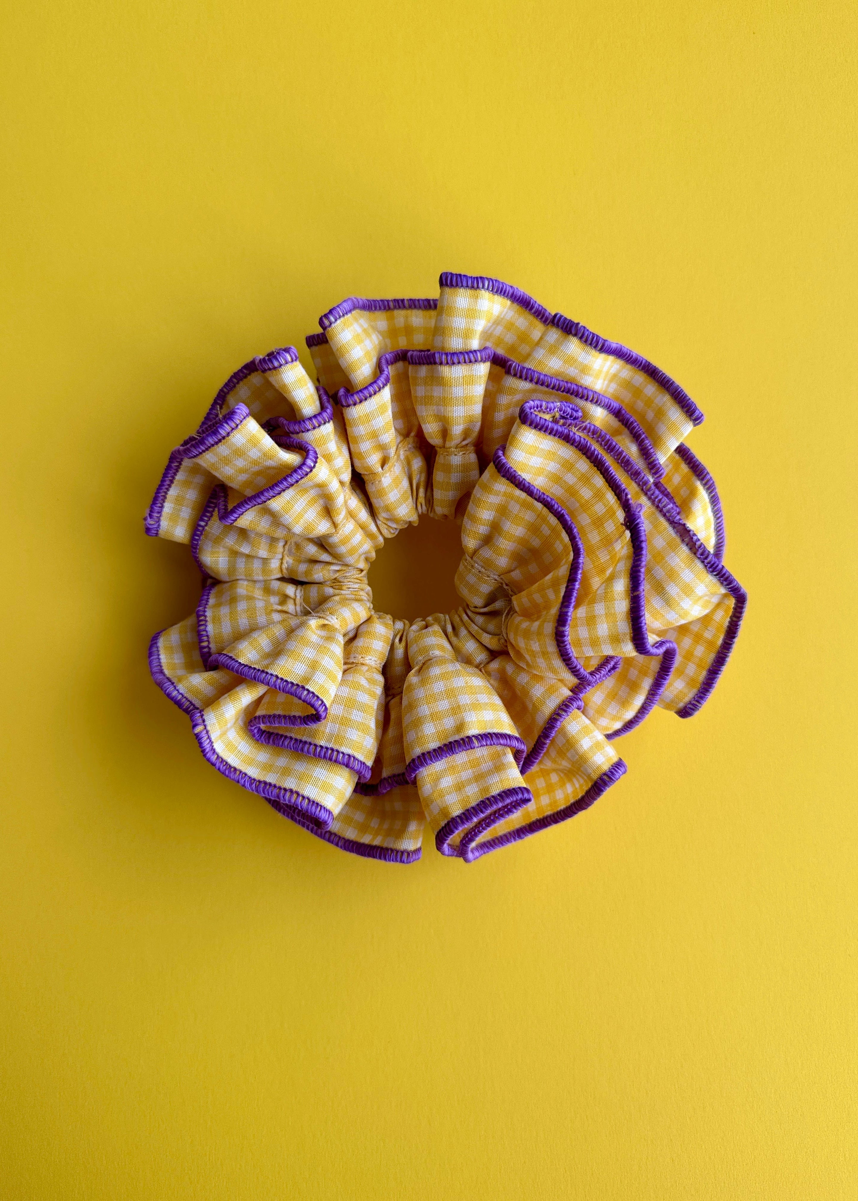 A ruffled fabric hair scrunchie in a yellow and white gingham pattern with purple trim, shown on a solid yellow background.