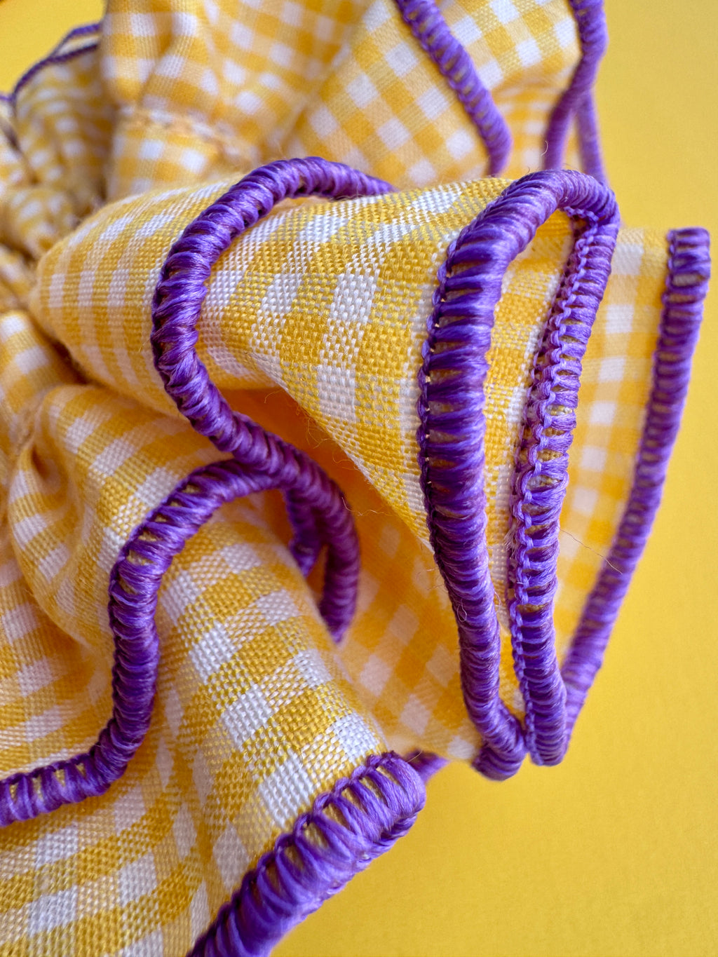 A close-up of a yellow and white gingham fabric item with purple braided trim, set against a solid yellow background.