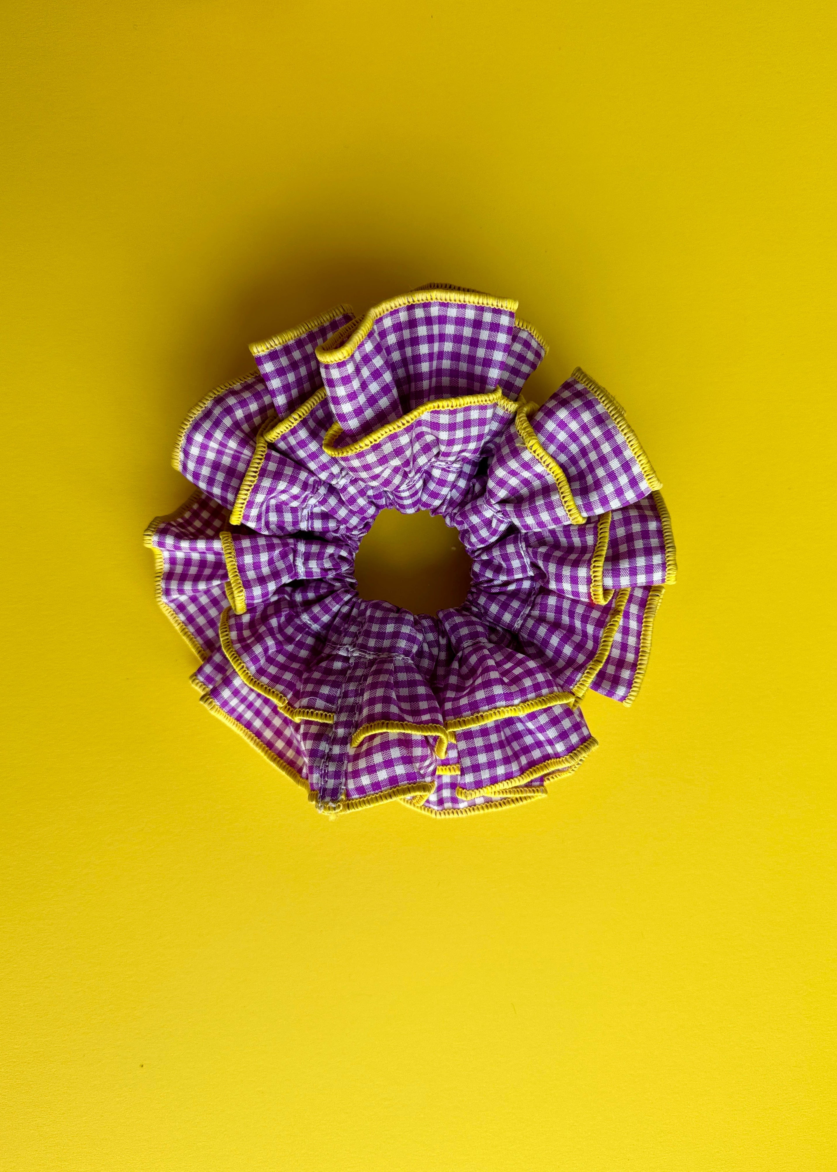 A fabric hair scrunchie with a purple and white gingham check pattern and yellow trim, photographed from directly above on a solid yellow background.