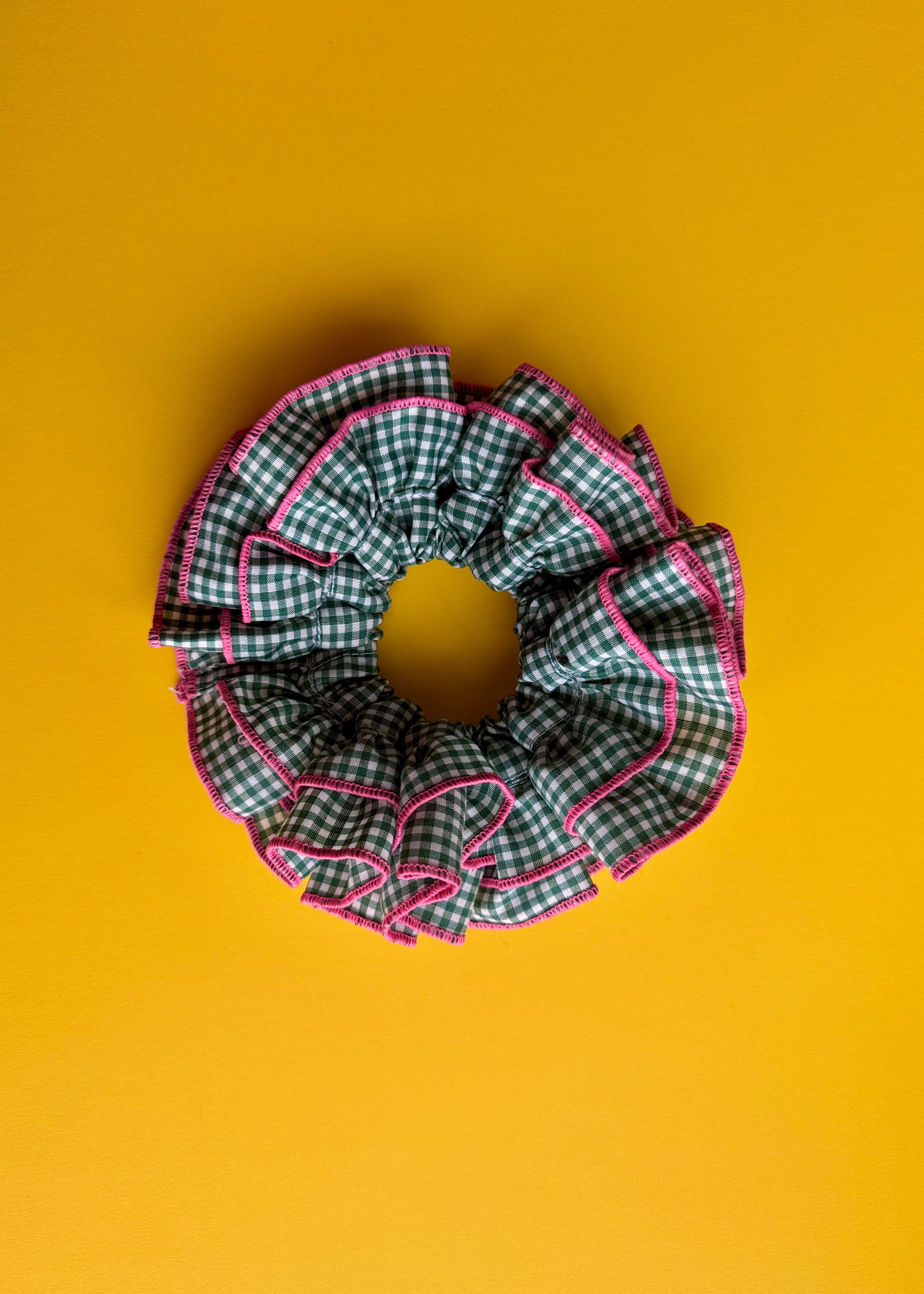 A single fabric hair scrunchie with a green and white gingham check pattern and bright pink piping, photographed from above on a solid mustard yellow background.
