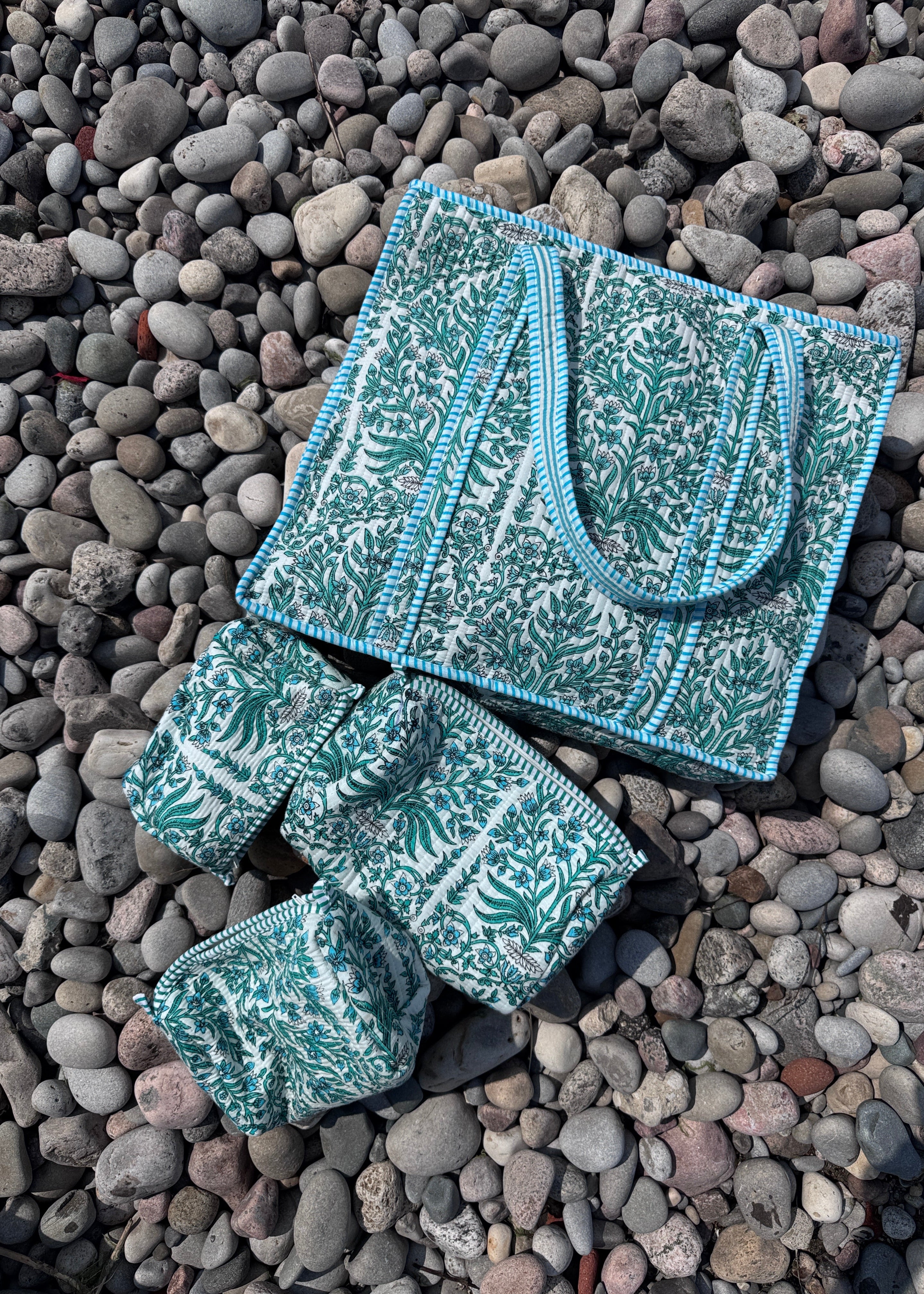 A matching set of three patterned fabric accessories, including a tote bag, a wristlet, and a small pouch, laid out on a bed of smooth gray pebbles. The fabric features a teal and white botanical pattern with light blue trim.