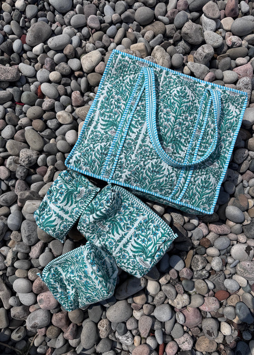 A matching set of three patterned fabric accessories, including a tote bag, a wristlet, and a small pouch, laid out on a bed of smooth gray pebbles. The fabric features a teal and white botanical pattern with light blue trim.